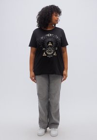 Černé tričko se zlatým grafickým motivem představujícím oko, slunce a nebeské prvky. Kombinováno se šedými kalhotami a bílými teniskami.