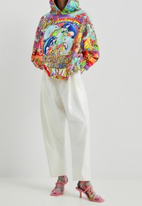 Hoodie coloré arborant un design vibrant de dauphin avec texte, associé à un pantalon blanc ample et des sandales à talons roses avec des accents décoratifs.