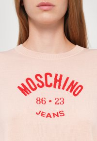 Pull léger rose avec un col rond côtelé. Présente un texte en rouge audacieux "MOSCHINO 86-23 JEANS" au design courbé. Texture de tissu douce.