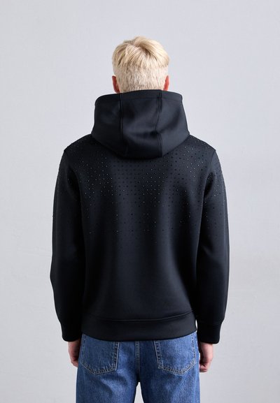 Hoodie preto com um capuz grande, apresentando um padrão texturizado de pequenos buracos nas costas. Combina com jeans de denim azul claro.