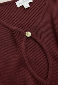 Bordeauxrotes, geripptes Umstandstop mit tiefem V-Ausschnitt und einem runden goldfarbenen Knopf in der Mitte, mit weicher Textur.