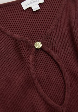 Burgundsko rebrast nosečniški top z globokim V-izrezom in okroglim zlatim gumbom na sredini, z mehko teksturo.