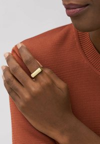 Goldfarbenen Metallring mit flachem rechteckigen Kopf. Wird an einer Hand getragen, die auf einem orangefarbenen, strukturierten Pullover ruht. Glatte Oberfläche und minimalistisches Design.