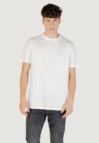 T-shirt bianco in cotone con maniche corte e scollatura rotonda. Presenta un piccolo logo rettangolare sulla parte sinistra del petto. Indossato con jeans in denim scuro.