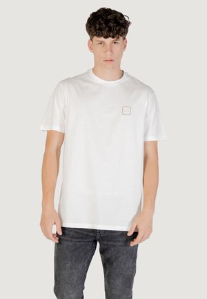 T-shirt bianco in cotone con maniche corte e scollatura rotonda. Presenta un piccolo logo rettangolare sulla parte sinistra del petto. Indossato con jeans in denim scuro.