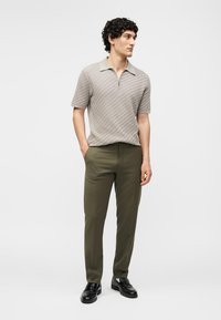 Άνδρας με μπεζ κοντομάνικο πόλο με υφή, ελαιδί παντελόνι και μαύρα επίσημα παπούτσια, στέκεται με τα χέρια στις τσέπες.