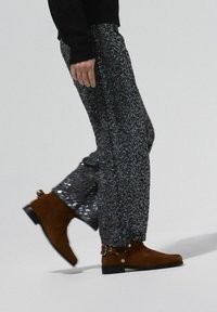 Pantalones brillantes de lentejuelas negras y plateadas combinados con botines marrones de gamuza con una correa y herrajes plateados, presentados en movimiento al caminar.