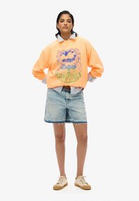 Sudadera oversized naranja con un diseño gráfico en azul y verde, combinada con shorts de mezclilla azul claro. Zapatillas beige completan el atuendo.