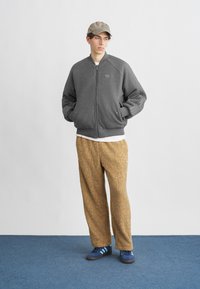 Tânăr îmbrăcat într-o jachetă gri cu fermoar, pantaloni bej pufoși, adidași albaștri și o șapcă bej, stând pe un covor albastru, în fața unui perete alb.