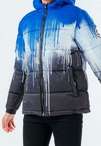 Chaqueta acolchada azul, blanca y negra con un patrón de goteo de pintura, cierre delantero, capucha y bolsillos laterales, llevada por una persona con pantalones negros.