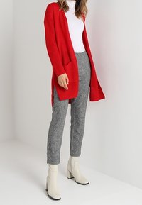 Cardigan rouge en tricot avec poches avant ouvertes, associé à un pantalon gris à motifs et des bottines blanches. Ensemble texturé et décontracté.