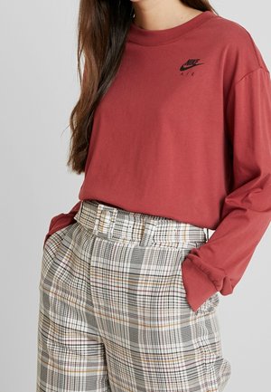 Femme portant un t-shirt rouge à manches longues Nike Air rentré dans un pantalon beige à carreaux taille haute avec une ceinture assortie, mains dans les poches.