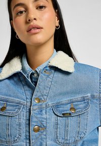 Chaqueta de mezclilla azul clara con un cuello de forro polar blanco, que presenta botones de bronce, dos bolsillos en el pecho y detalles de costura visibles.