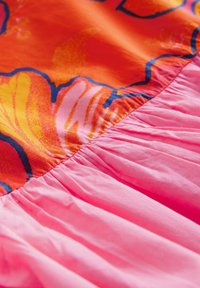 Blommönstrad tyg i orange och gult övergår i en rosa, draperad del. Materialen verkar släta med en lätt glans.