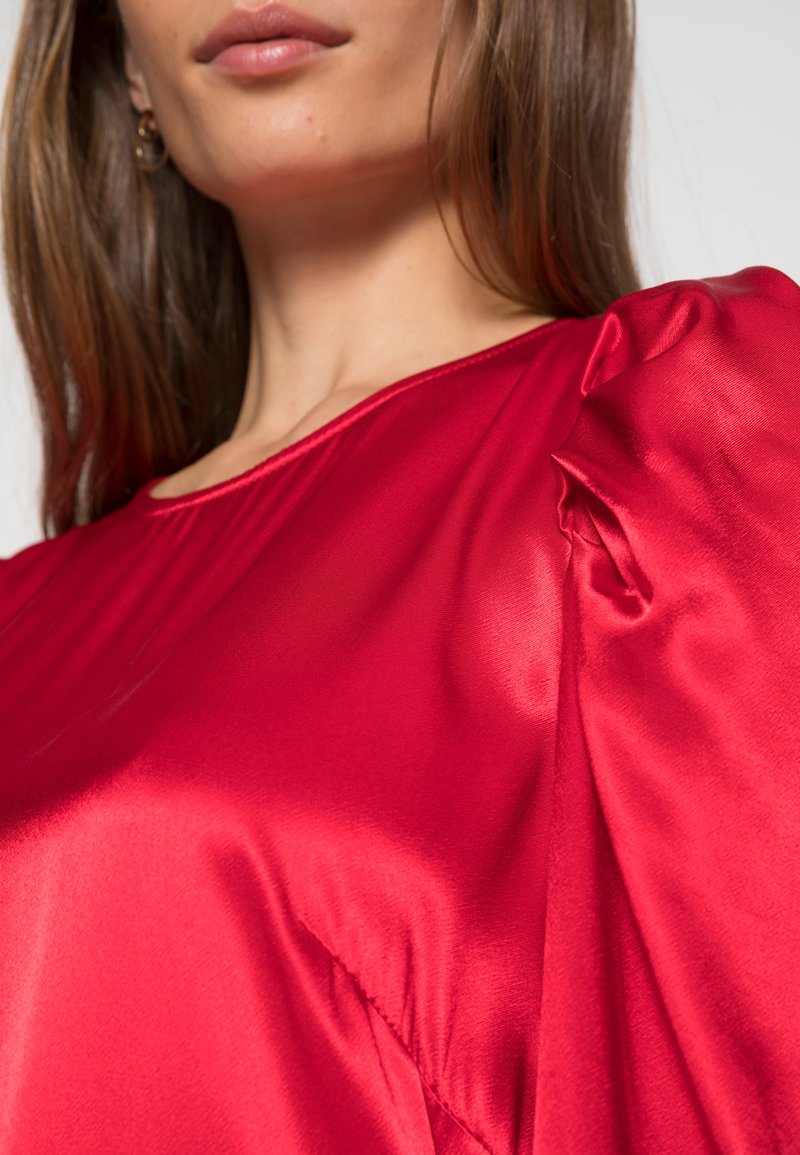 Rotes Satin-Oberteil mit einem tiefen Ausschnitt und puffärmel, das eine glatte, glänzende Textur und ein drapiertes Design nahe der Schulter zeigt.