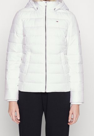 Personne portant une veste matelassée blanche zippée à capuche avec poches latérales et un pantalon noir, debout devant un fond blanc uni.