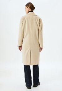 Beige uldfrakke med lige snit, lange ærmer og klapkrave. Har en bagventil og minimale syninger. Bæres over navy jeans.