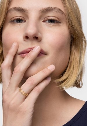 Goldring mit ineinander verschlungenen herzförmigen Gliedern, getragen an einer Hand mit natürlichen Nägeln; sanfte, neutral getönte Haut; kurzes, hellblondes Haar.