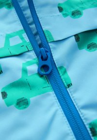 Světle modrá látka s vzorem zelených náklaďáků, uprostřed se nachází modrý zip. Je viditelná hladká textura a detaily švu.
