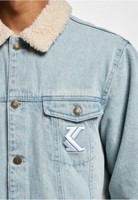 Chaqueta de denim azul claro con cuello de sherpa crema, botones de metal y logotipo bordado en blanco sobre el bolsillo izquierdo del pecho.