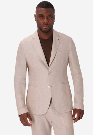 Uomo che indossa una giacca da completo beige chiaro e pantaloni abbinati su una camicia marrone scuro, in piedi davanti a uno sfondo bianco semplice.