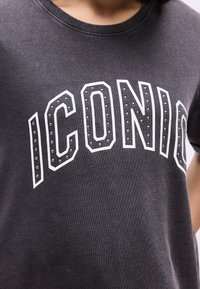 Κοντινό πλάνο μαύρου μπλουζακιού με μεγάλες άσπρες γράμμες "ICONIC", περιγραμμένες και διακοσμημένες με μικρά μεταλλικά καρφιά στο στήθος.