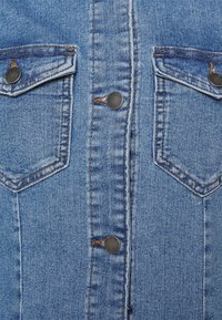 Denimjacka med ljusblått tyg, två bröstfickor med mörka metallknappar och synliga sömmar längs kanterna.