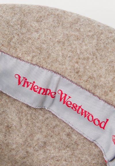Beige vilnas materiāls ar gludu tekstūru; tam ir sudraba lente ar sarkano "Vivienne Westwood" logo, kas izšūta, demonstrējot zīmola identitāti.