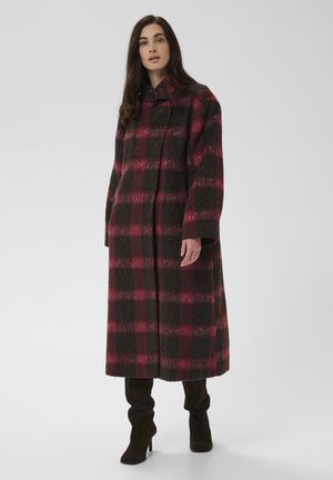 Femme portant un long manteau à carreaux rouges et noirs avec un col haut et des bottes noires à talons, debout sur un fond uni.
