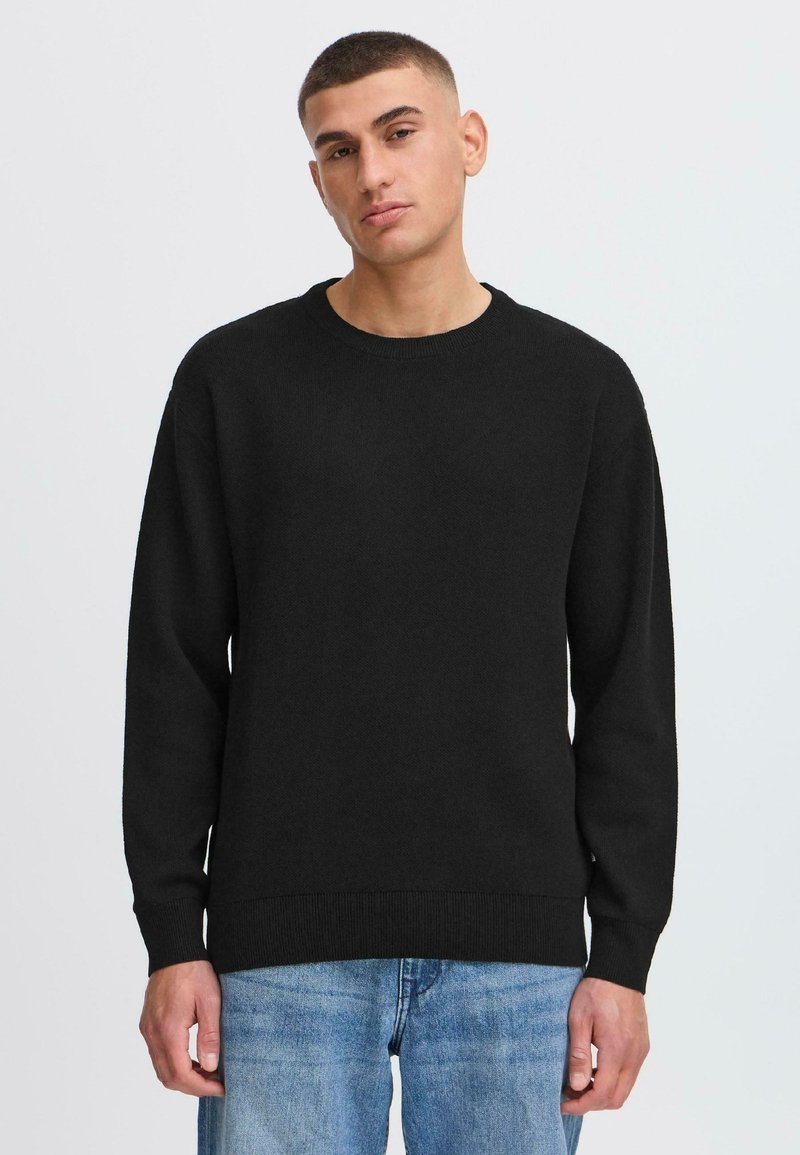 Svart strikkegenser med rund hals og lange ermer. Har ribbestrikkede detaljer på mansjettene og kanten, bæres over blå slitte jeans.
