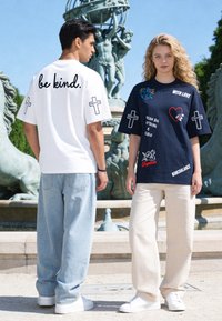 Jeune homme en t-shirt blanc « be kind. » et femme en t-shirt graphique bleu marine se tiennent près d'une fontaine avec des statues sous un ciel dégagé.