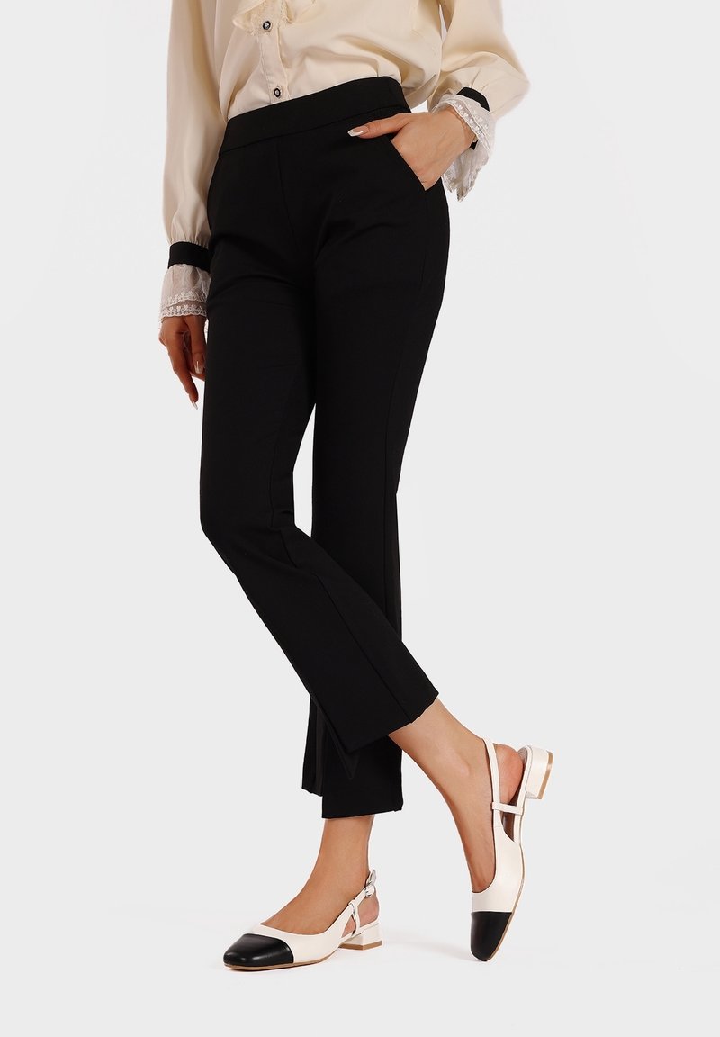 Femme portant un pantalon slim noir et un chemisier beige avec des poignets en dentelle, debout avec une main dans la poche, portant des chaussures plates beige et noires.