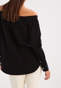 Svart axelbandslös skjorta med elastisk halsringning, långa ärmar och knäppta manschetter. Mjukt tyg, rundad fåll och slät textur.