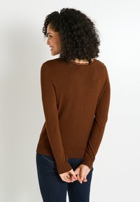 Femme avec des cheveux bouclés portant un pull marron à manches longues et un jean bleu foncé, se tenant avec les mains jointes derrière le dos.