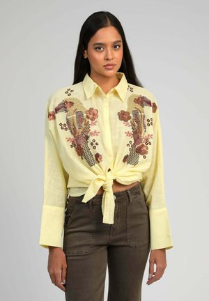 Femme aux longs cheveux bruns portant une chemise boutonnée jaune clair nouée à la taille, ornée de broderies colorées d'oiseaux et de fleurs, assortie à un pantalon foncé.