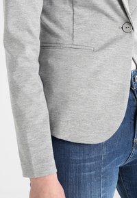 Gros plan sur une personne portant un blazer gris clair avec des détails de boutons et de poches, accompagné d’un jean bleu en denim.