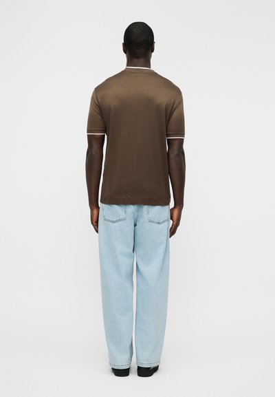 Καφέ κοντομάνικη μπλούζα με λευκή επένδυση, σε συνδυασμό με ανοιχτό μπλε τζιν παντελόνι με φαρδιές μπατζάκια. Ορατή ραφή στην πλάτη και δύο πίσω τσέπες στο τζιν.