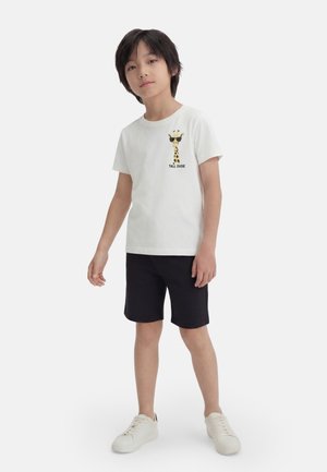 Dreng iført hvid t-shirt med giraf-grafik og teksten "Tall Dude", sorte shorts og hvide sneakers, stående foran en ensfarvet baggrund.