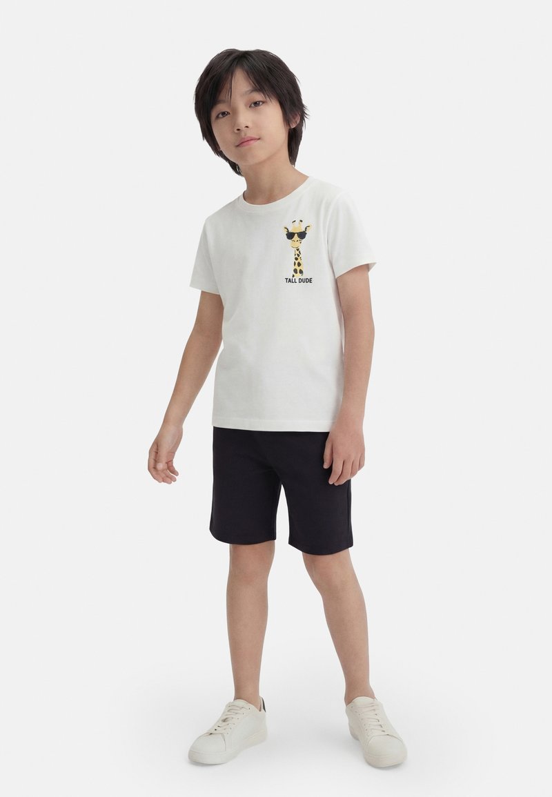 Garçon portant un t-shirt blanc avec un dessin de girafe et le texte « Tall Dude », un short noir et des baskets blanches, debout sur un fond uni.