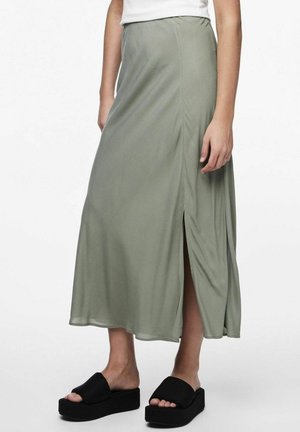 Jupe maxi vert olive avec une fente sur le côté, fabriquée en tissu lisse. Portée avec des sandales à plateforme noires. Design simple et fluide.