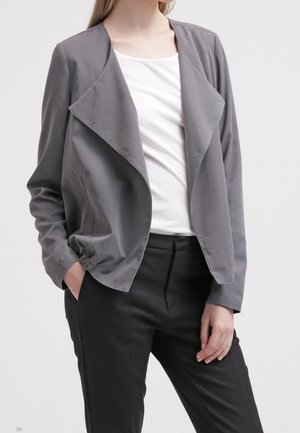 Kvinde iført en løs grå åben-front blazer, hvid top og sorte bukser med den ene hånd i lommen mod en ensfarvet baggrund.