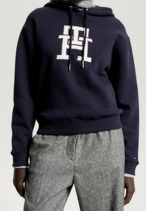Personne portant un sweat à capuche bleu marine avec un logo blanc brodé et un pantalon en laine gris sur un fond uni.