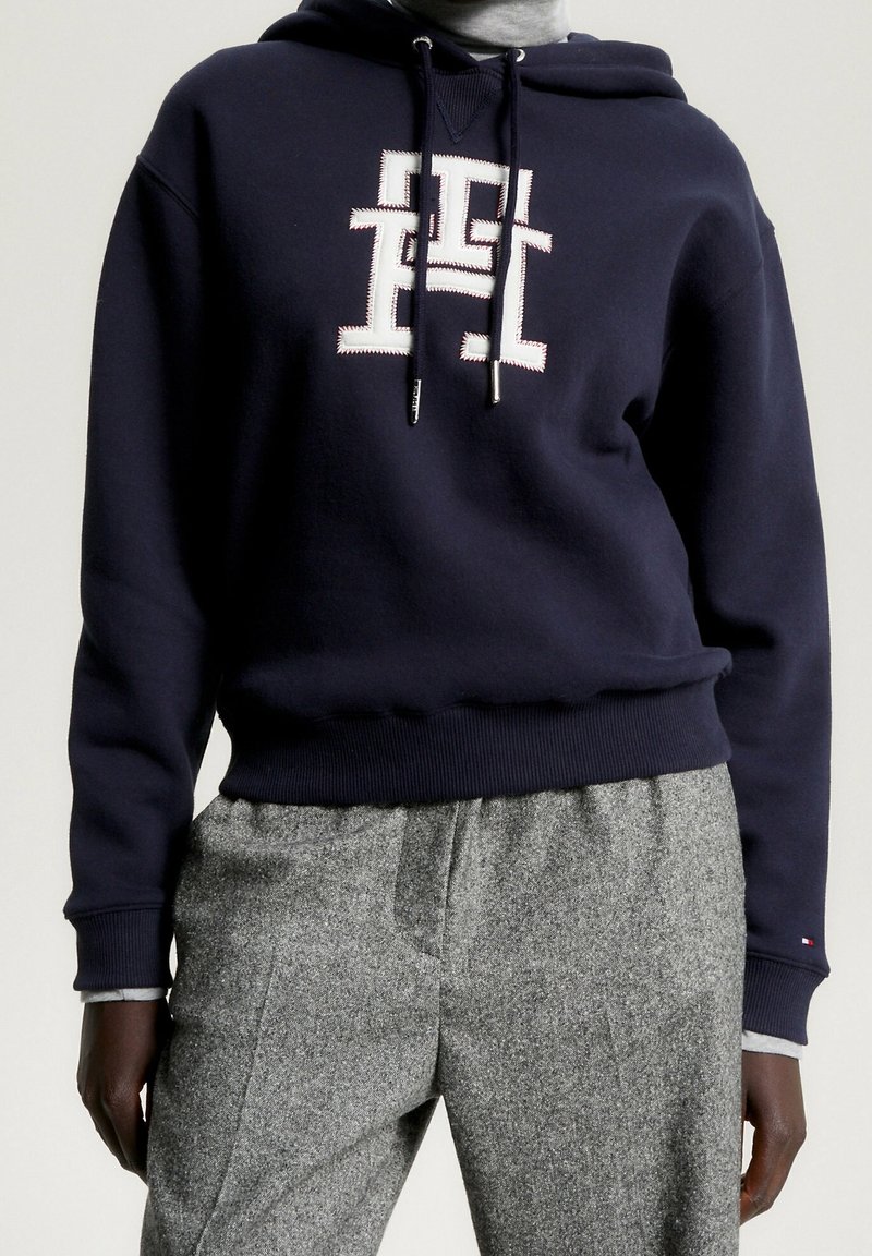 Personne portant un sweat à capuche bleu marine avec un logo blanc brodé et un pantalon en laine gris sur un fond uni.