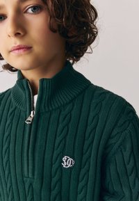 Jersey de punto verde oscuro con cuello alto y cremallera plateada. Presenta un patrón de punto trenzado y un pequeño logo bordado en el pecho.
