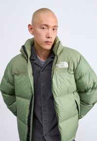 Mees, kellel on raseeritud pea, kannab rohelist The North Face'i puhversjakki musta nööpide jaoks särke, vaatab kaamerasse rahulikult, taust on neutraalne.