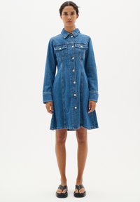 Robe en jean avec un col, des manches longues, une fermeture à boutons sur le devant et deux poches poitrine. Confectionnée en denim bleu moyen avec une jupe évasée.