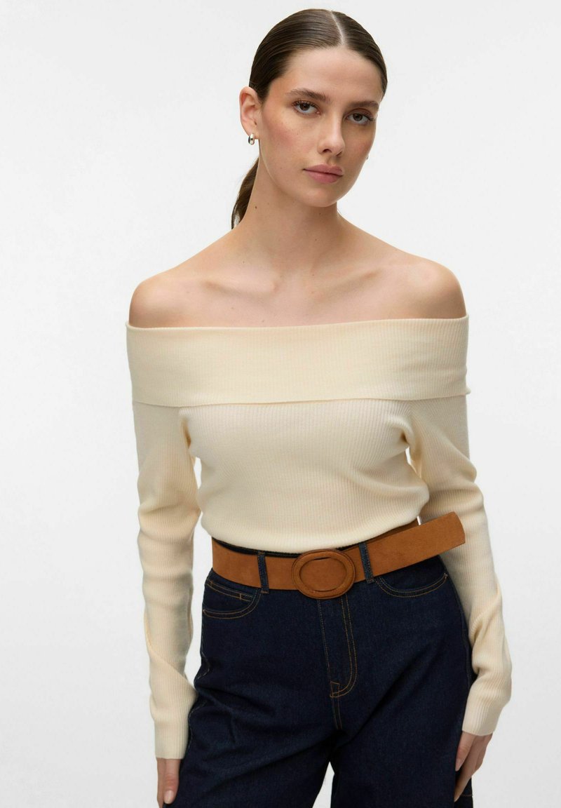 Femme aux cheveux tirés en arrière, portant un pull crème à épaules dénudées, un jean taille haute foncé et une ceinture marron avec une boucle ronde, debout devant un fond blanc.