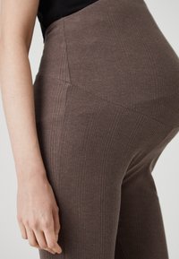 Bruna ribbade leggings med hög midja och en slät yta. Närbild visar sid sömn detaljer och en hand nära midjan.