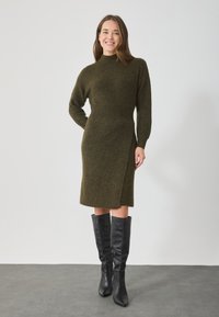 Kötött olívazöld ruha magas gallérral, hosszú ujjakkal és aszimmetrikus szegéllyel, térd felett érő fekete bőr csizmával párosítva.