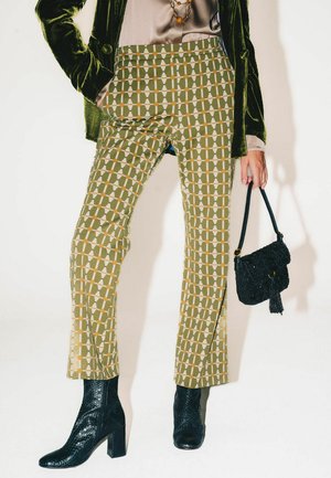 Grün gemusterte Hosen mit geometrischen Formen in Orange und Weiß, kombiniert mit schwarzen Stiefeletten und einer kleinen schwarzen Handtasche mit Quaste.